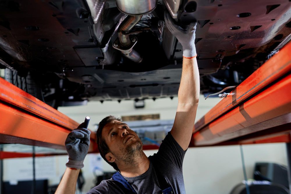 Mechanic working under a car on a lift, using a flashlight, wearing gloves.