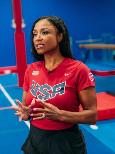 Dominique Dawes in red USA shirt speaking at her gymnastic studio