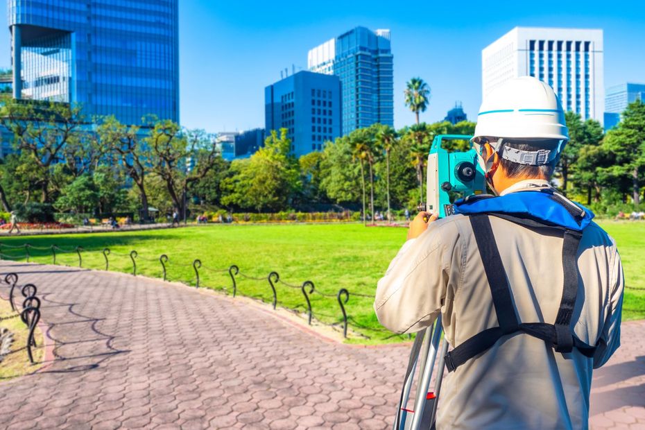 Worker surveying a city park with a tripod, facing modern glass buildings and green lawns