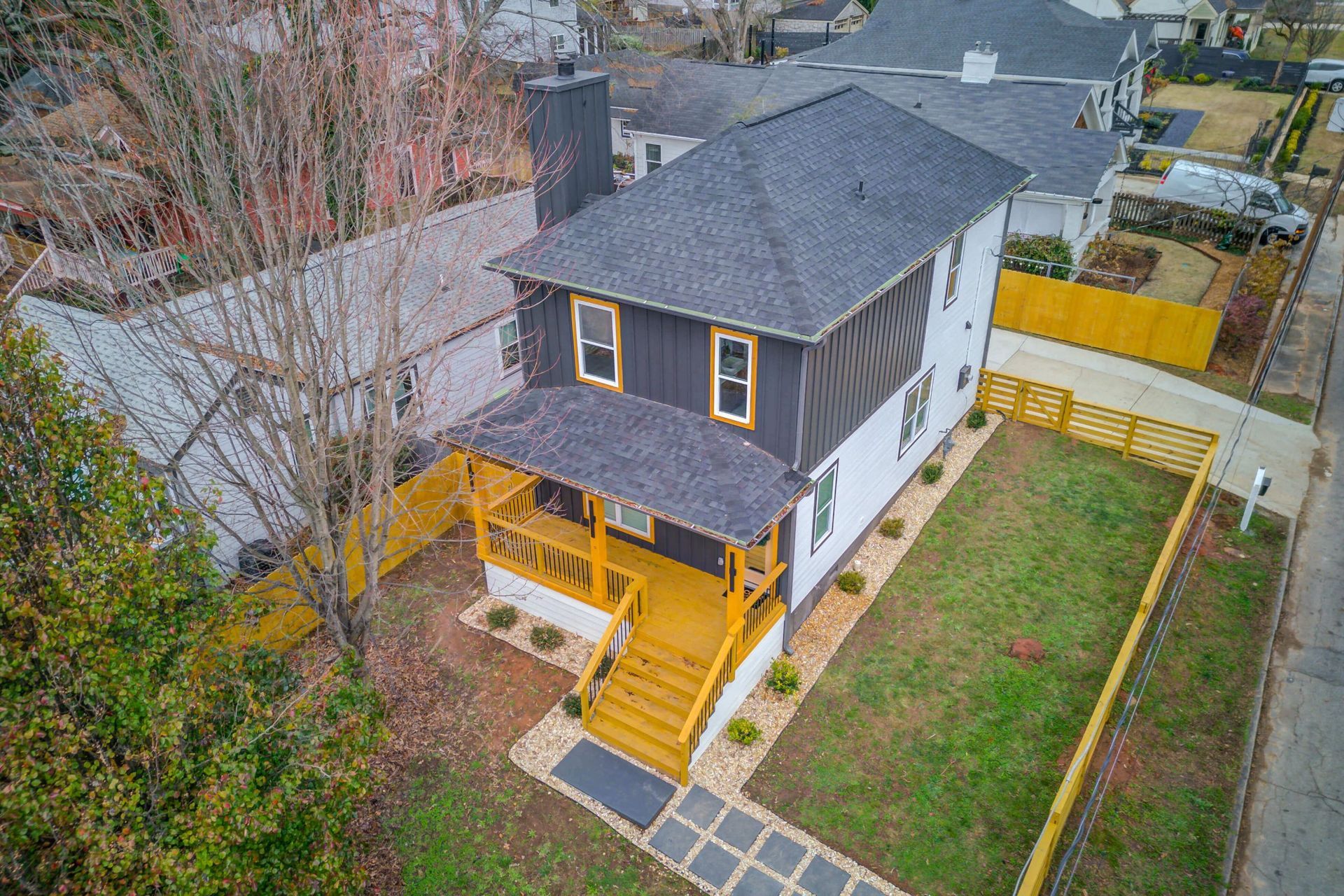 An aerial view of a two-story gray house with a yellow porch and steps, surrounded by a yellow fence and a grassy yard.