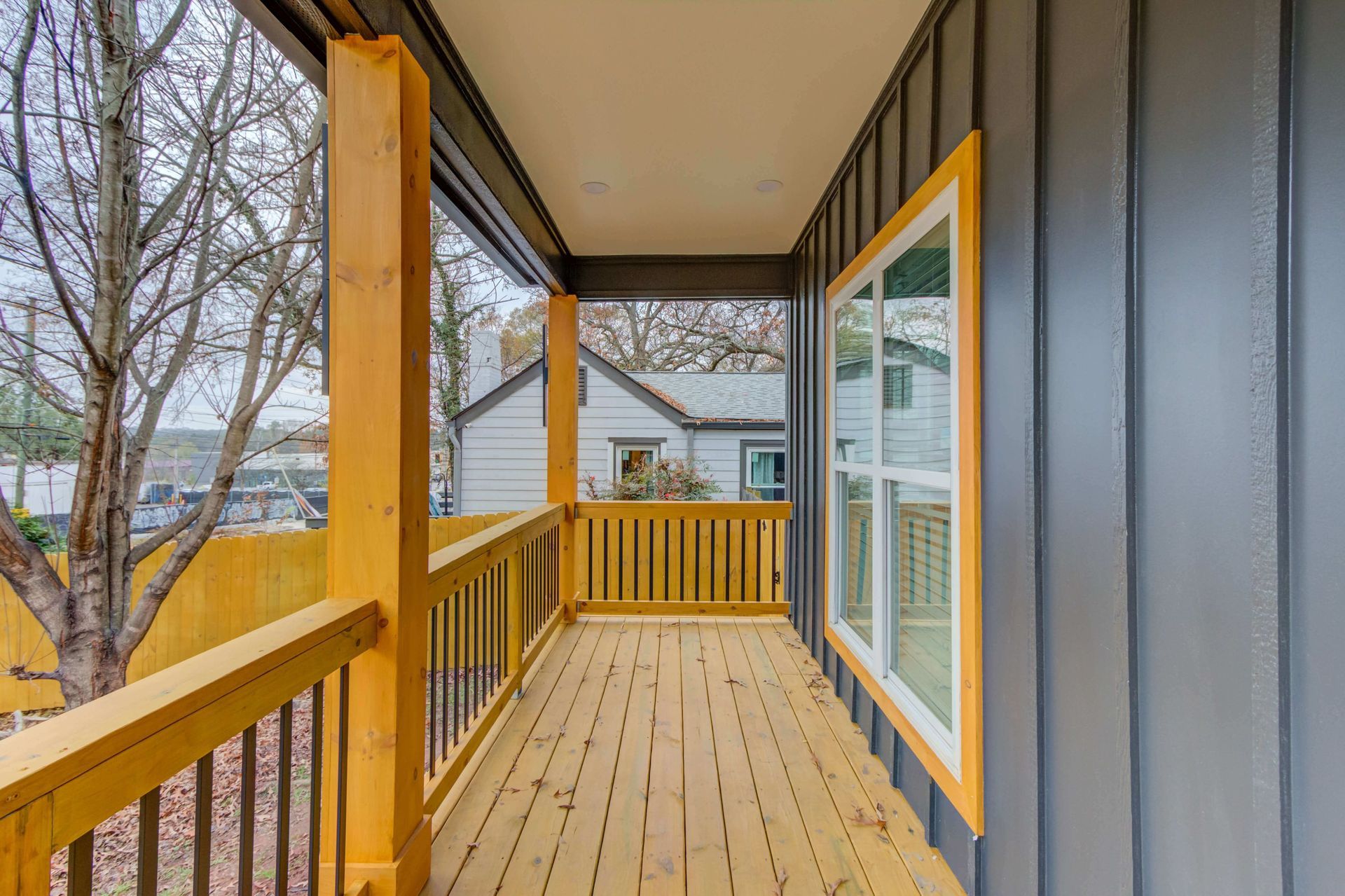 A covered wooden porch with a yellow railing and trim against grey vertical siding, overlooking a fenced outdoor area.