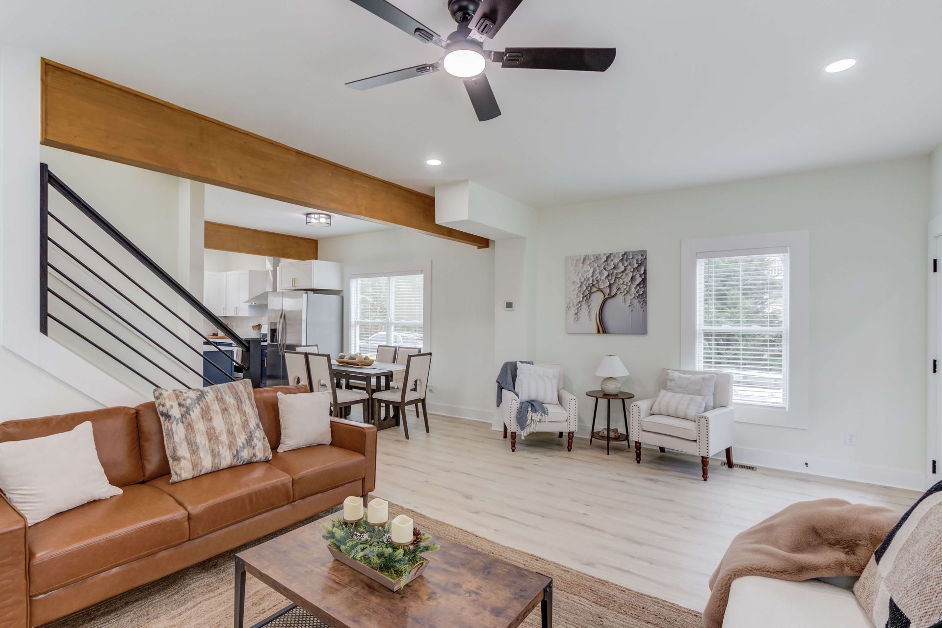 A modern living room with a brown leather sofa, white armchairs, a wooden coffee table, and an open dining area.
