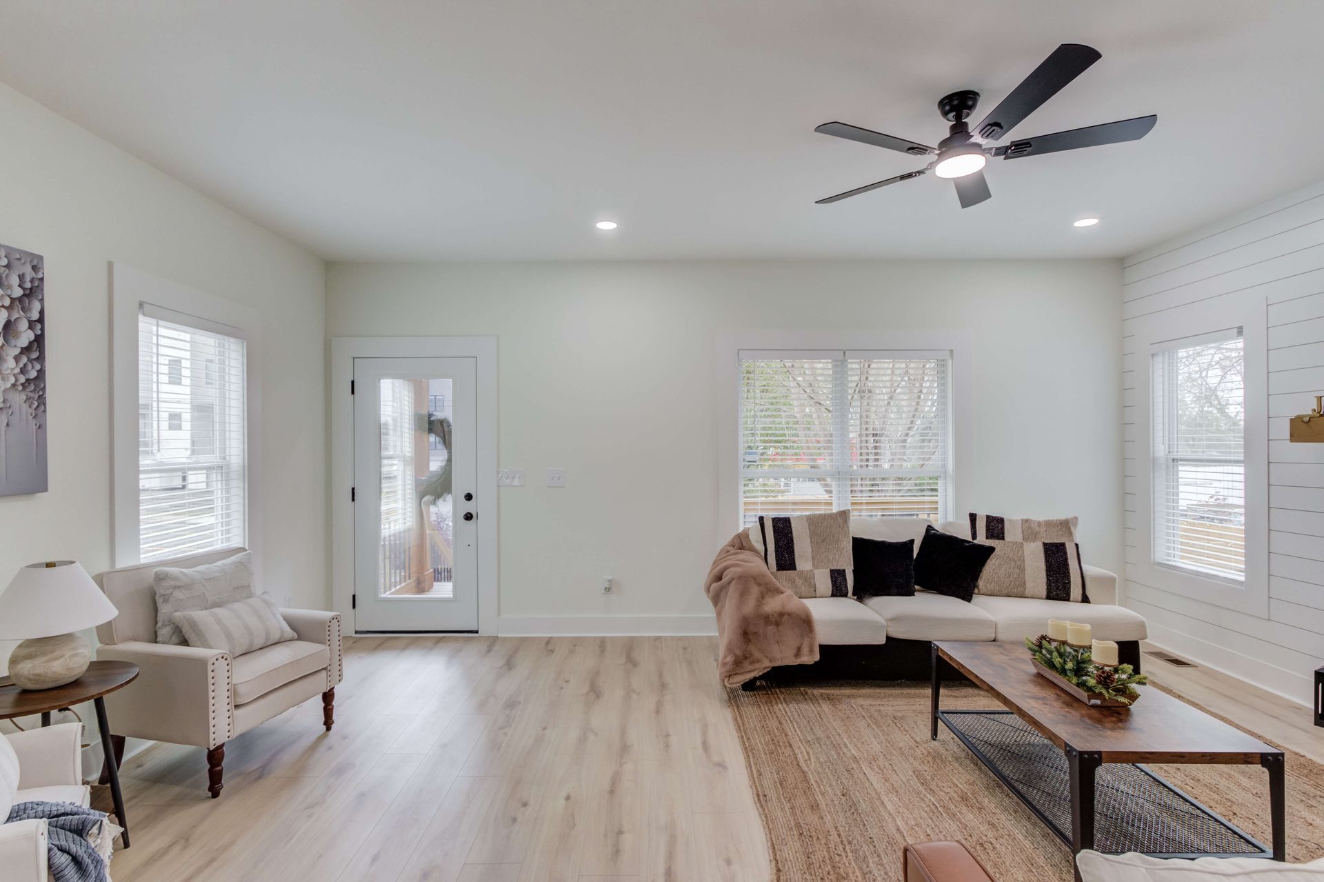 A bright living room with light wood floors, a beige sofa with dark pillows, an armchair, and a wood-frame coffee table.
