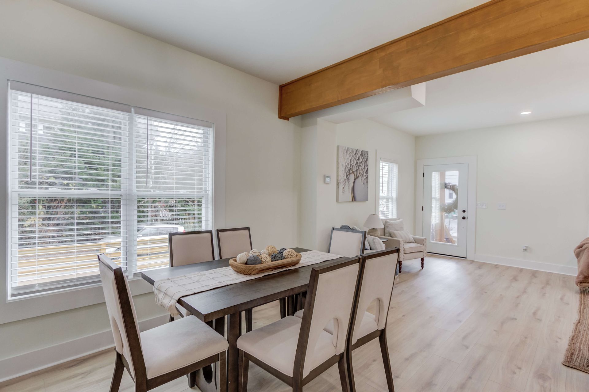 A dining area with a wood-toned beam, a table with four beige chairs, light wood floors, and a bright white entryway.