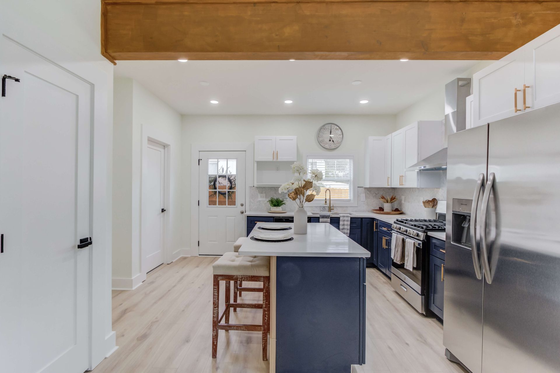 A bright, modern kitchen featuring a dark blue island with stools, white cabinets, stainless appliances, and wood flooring.