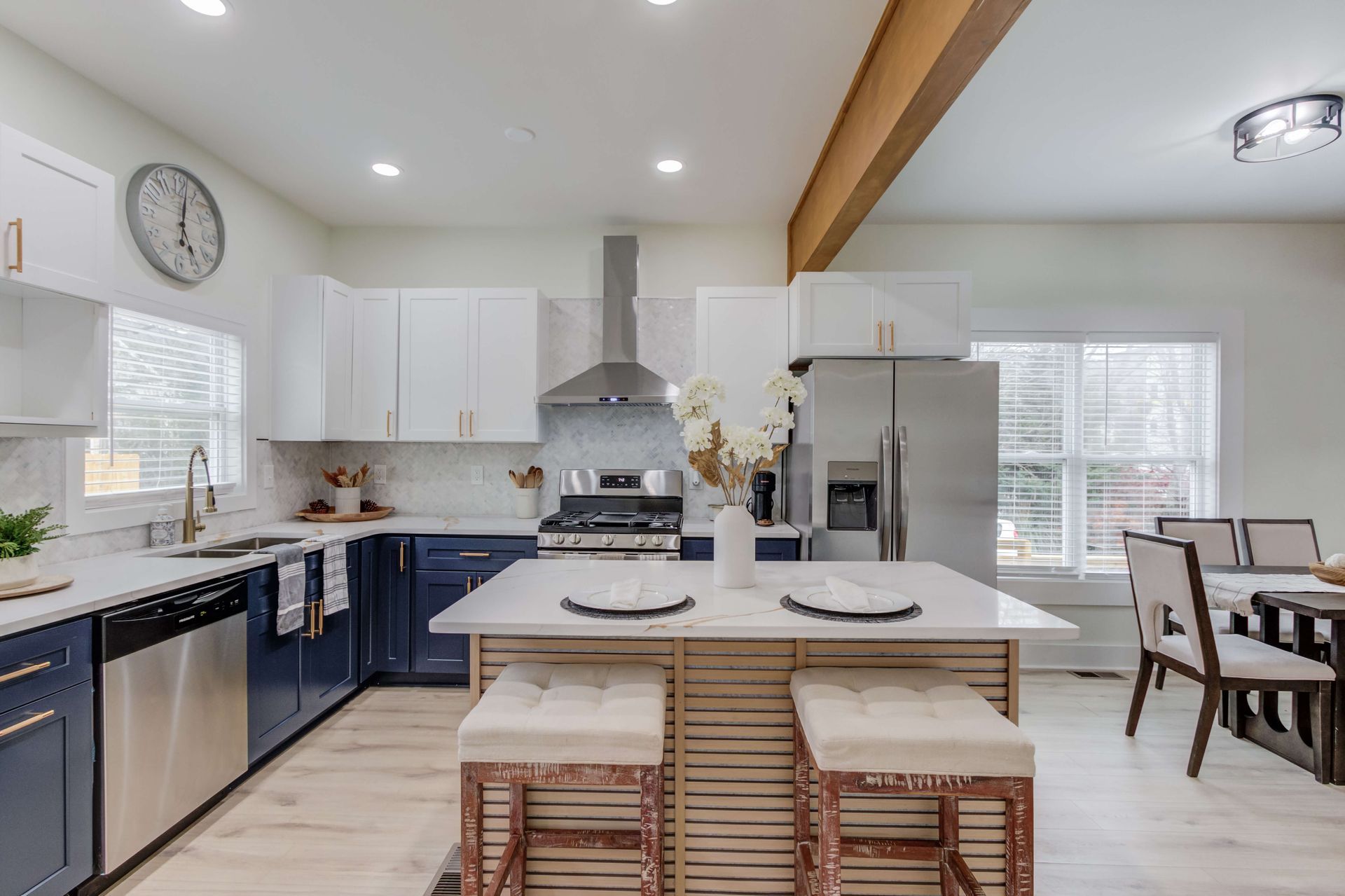 A modern kitchen with white upper cabinets, dark blue lower cabinets, a central island with stools, and stainless appliances.
