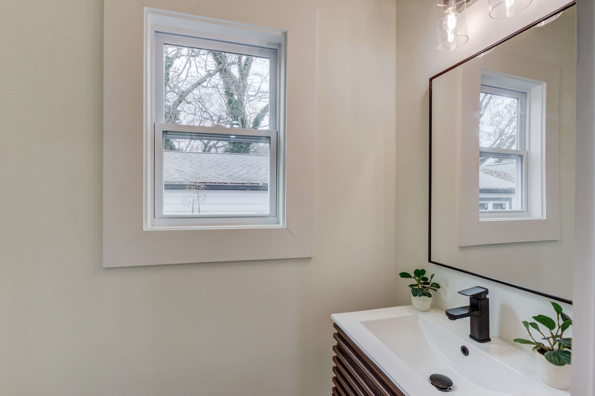 A bathroom sink vanity with a black faucet, small potted plants, and a framed mirror reflecting a window in a bright room.