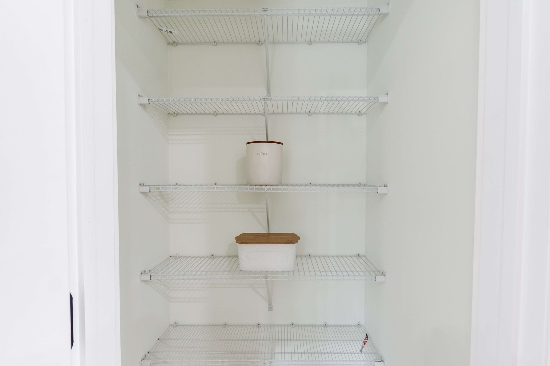 A white closet with four wire shelves; one shelf holds a small canister, and another holds a rectangular storage container.