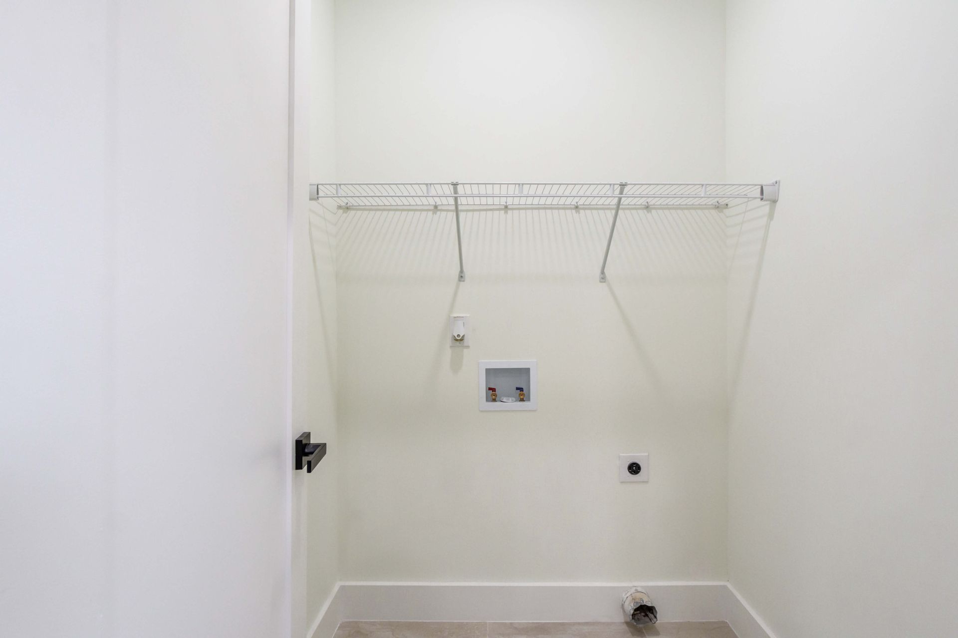 A small, white laundry closet containing a wall-mounted wire shelf, utility connections, and a drain outlet.