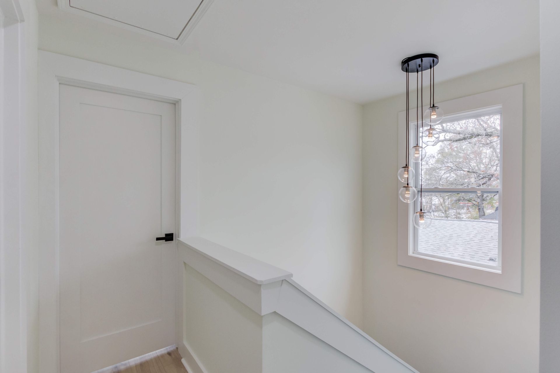 A white hallway with a door, a staircase railing, and a modern pendant light fixture hanging near a window.