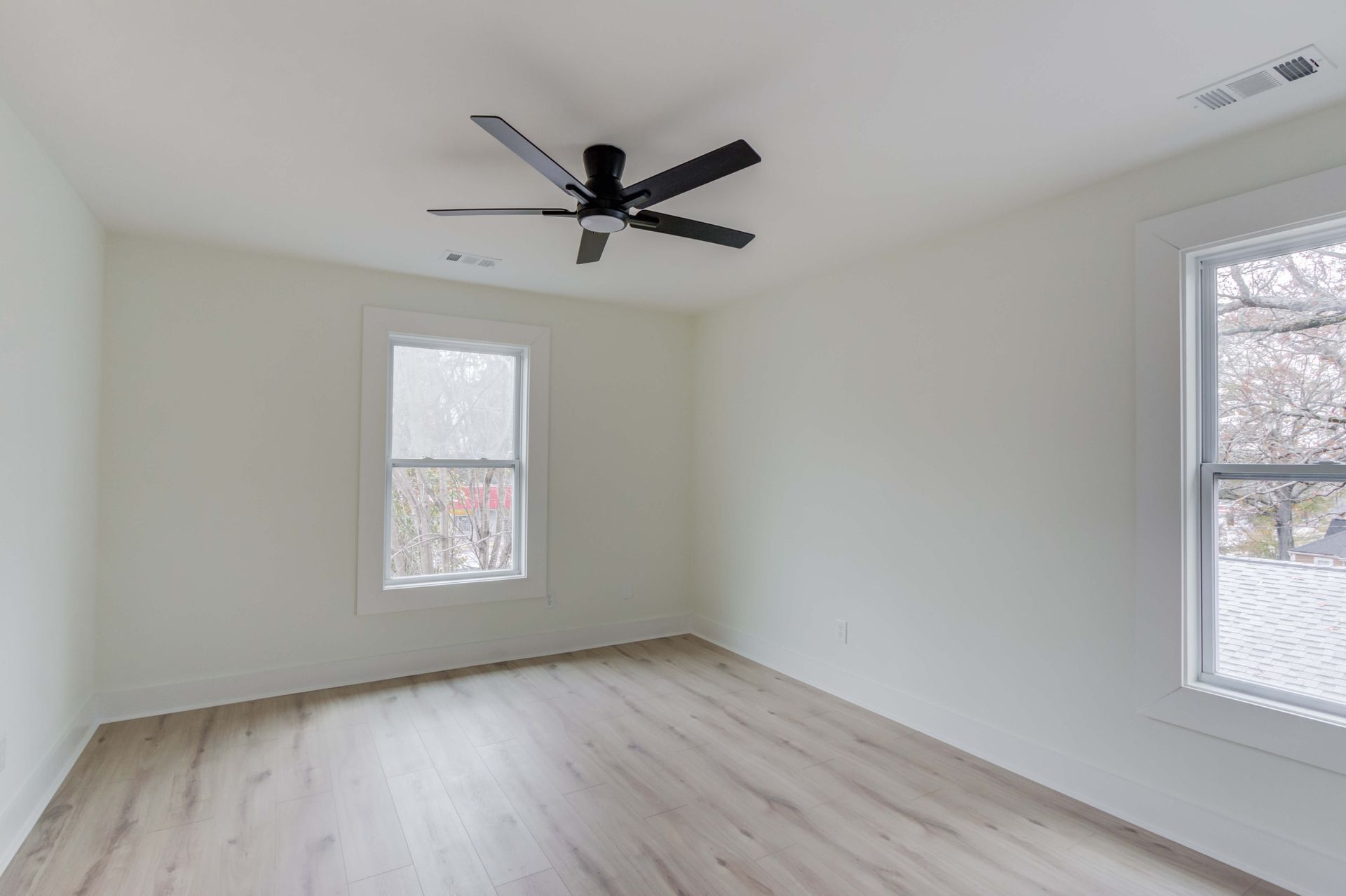 An empty room with light wood-look flooring, white walls, two windows, and a black ceiling fan.