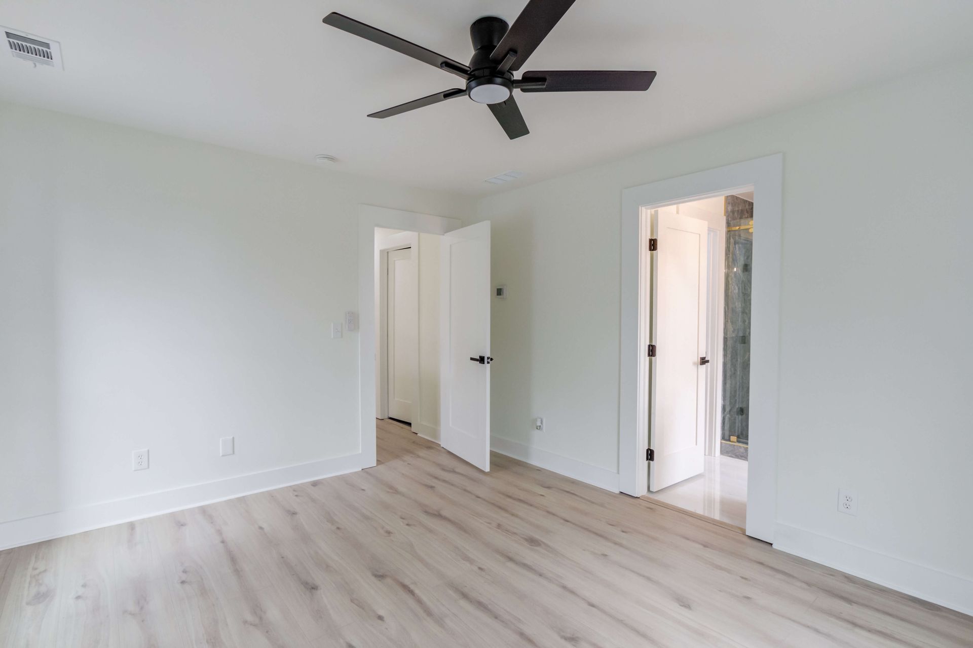 An empty room with light wood-look floors, white walls, a ceiling fan, and two open doorways leading to other rooms.