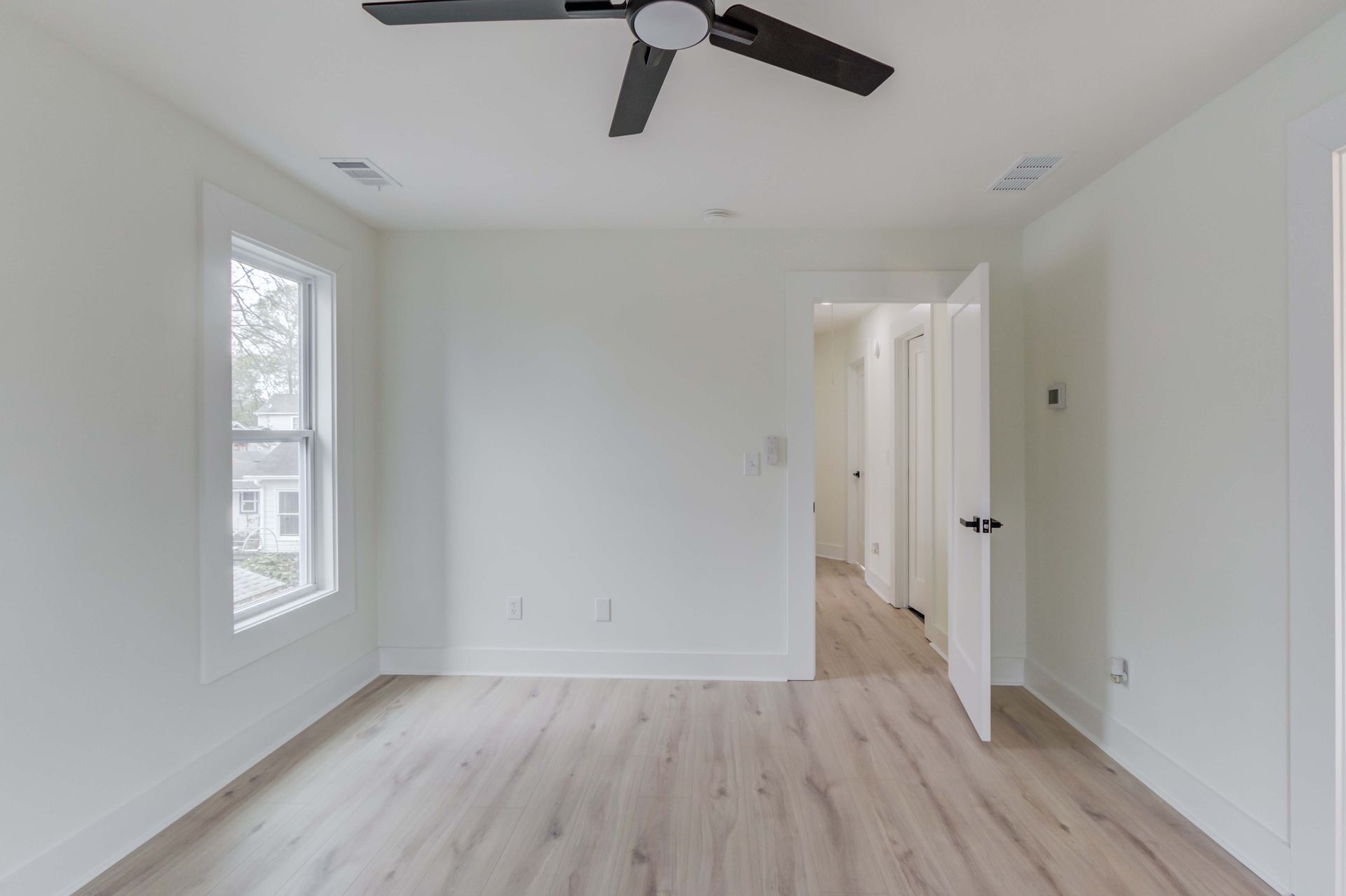A minimalist room with white walls, light wood flooring, a window, a ceiling fan, and an open doorway leading to a hallway.
