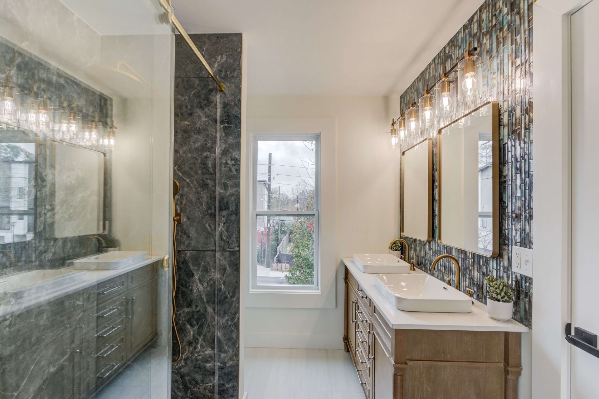 A modern bathroom with two marble-topped vanities, gold fixtures, and a large window set against a dark marble wall.