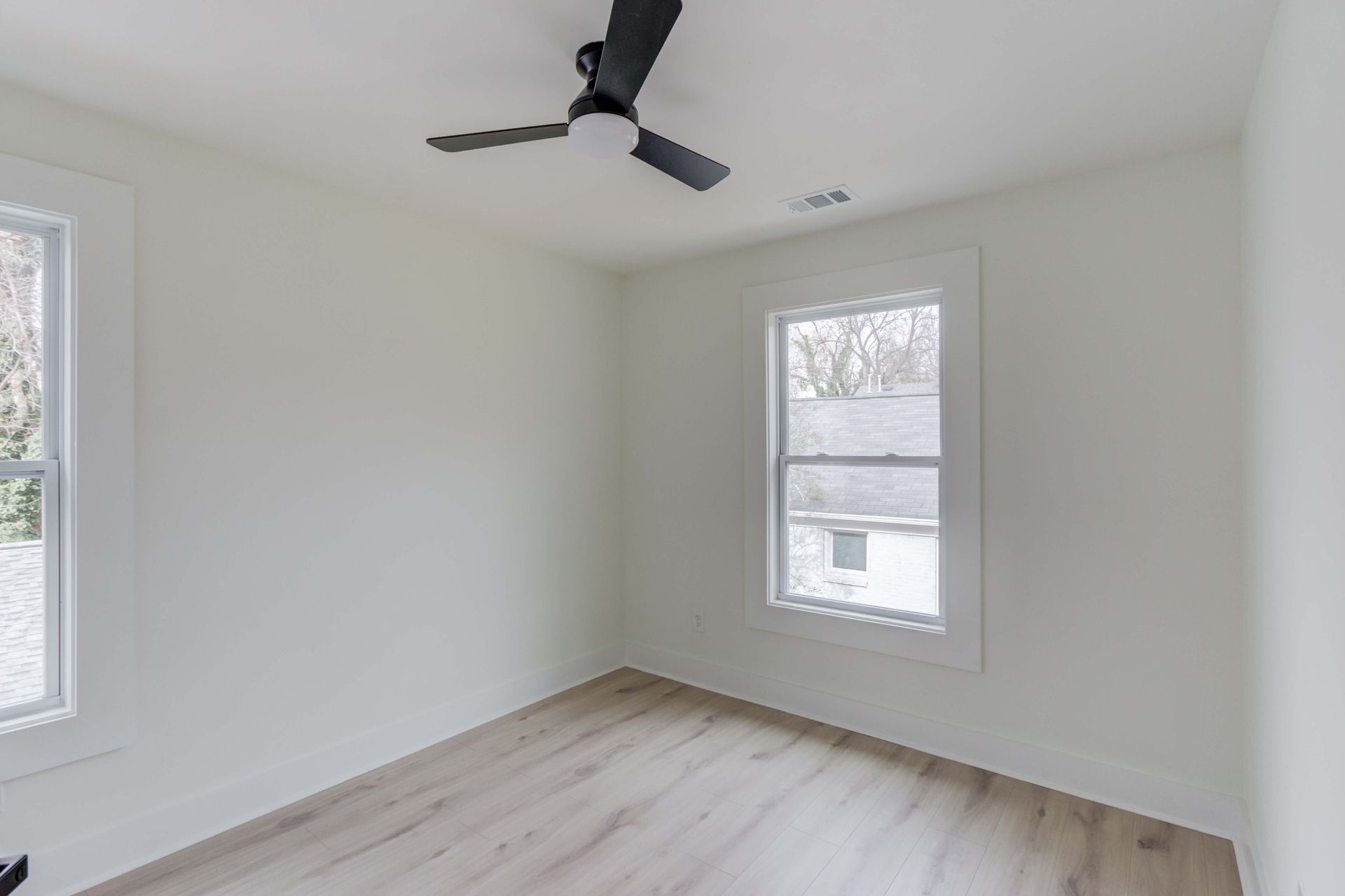 An empty room with white walls, light wood-look flooring, two windows, and a black ceiling fan.