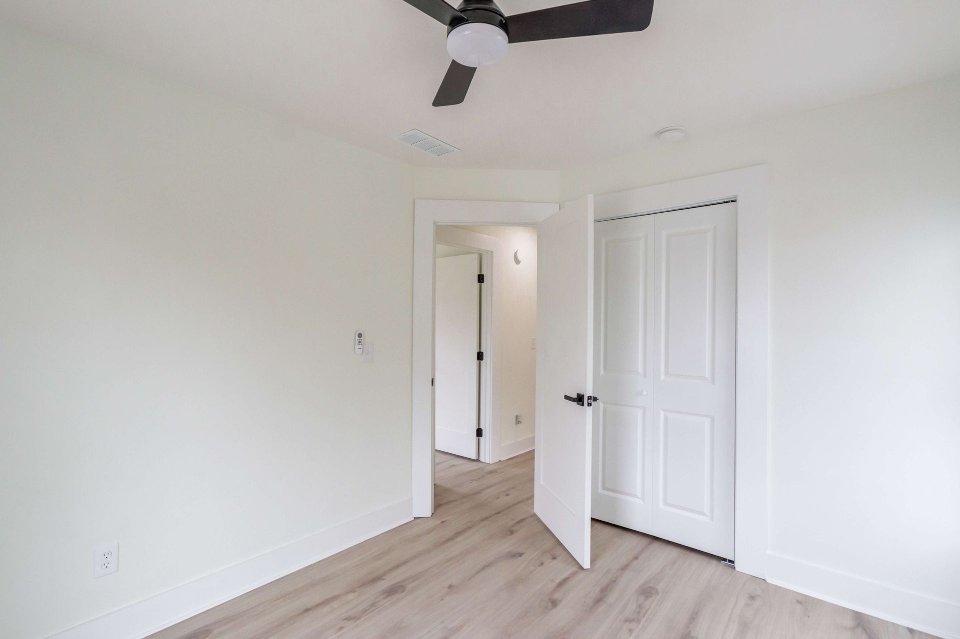 A brightly lit room with white walls, light wood flooring, a ceiling fan, and open doorways leading to other spaces.