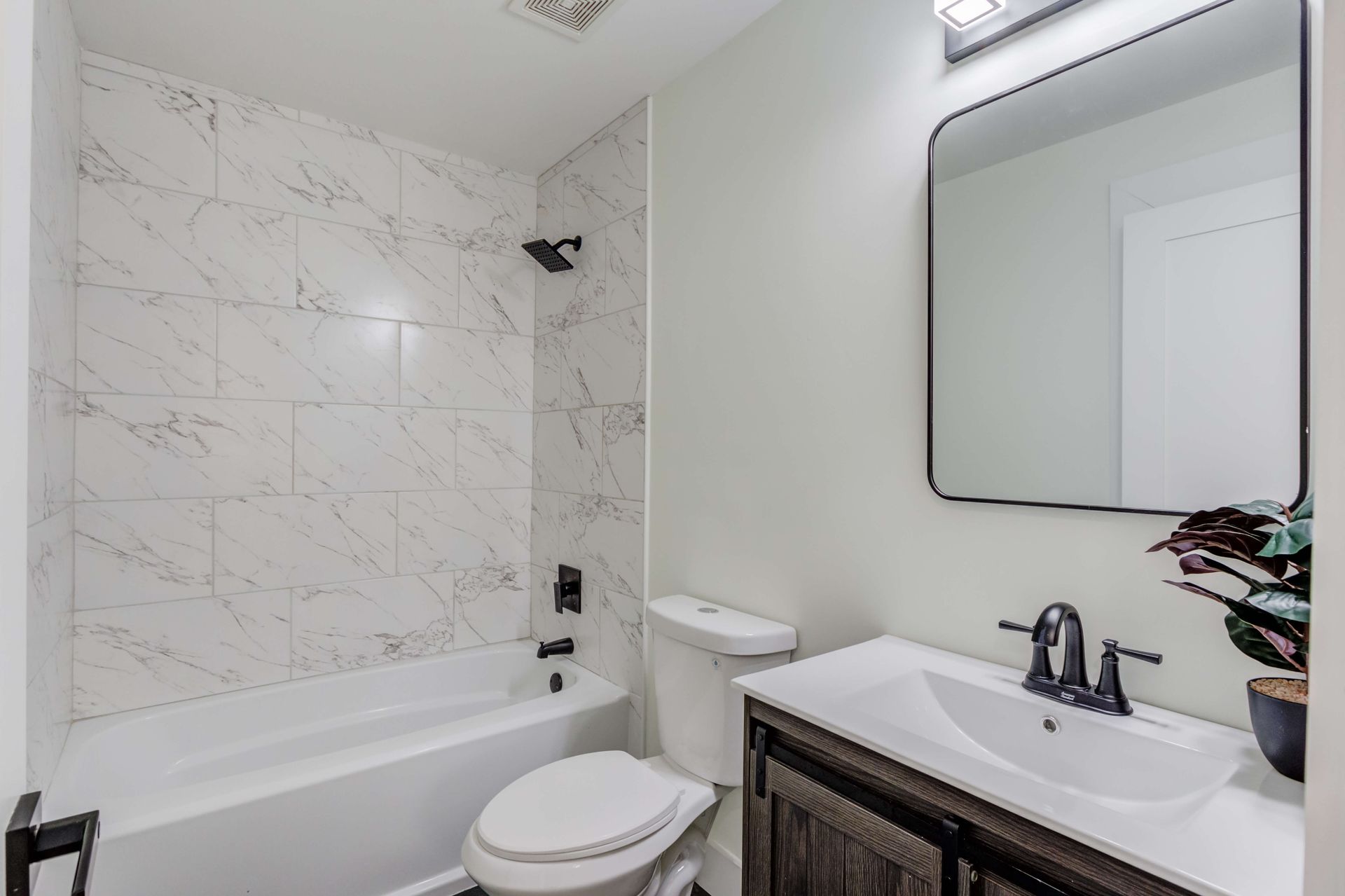A modern bathroom featuring a white bathtub with marble-style tile walls, a toilet, and a vanity with a black faucet.