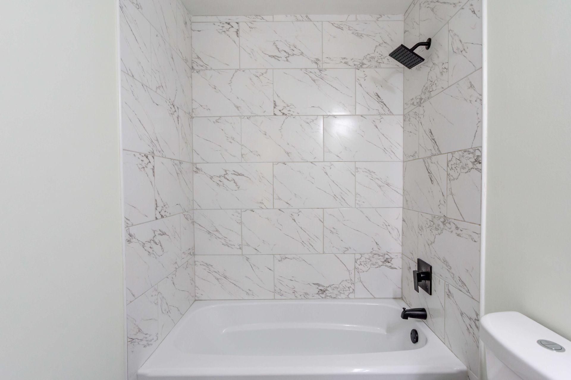 A white bathtub and shower surround with marble-look rectangular wall tiles and matte black fixtures in a bathroom.