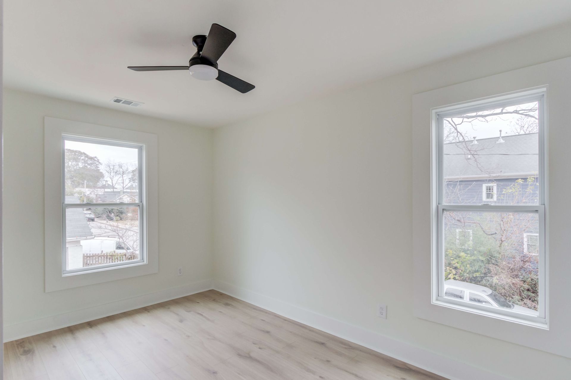 Empty room with off-white walls, wood-look flooring, a modern black ceiling fan, and two windows facing outdoors.