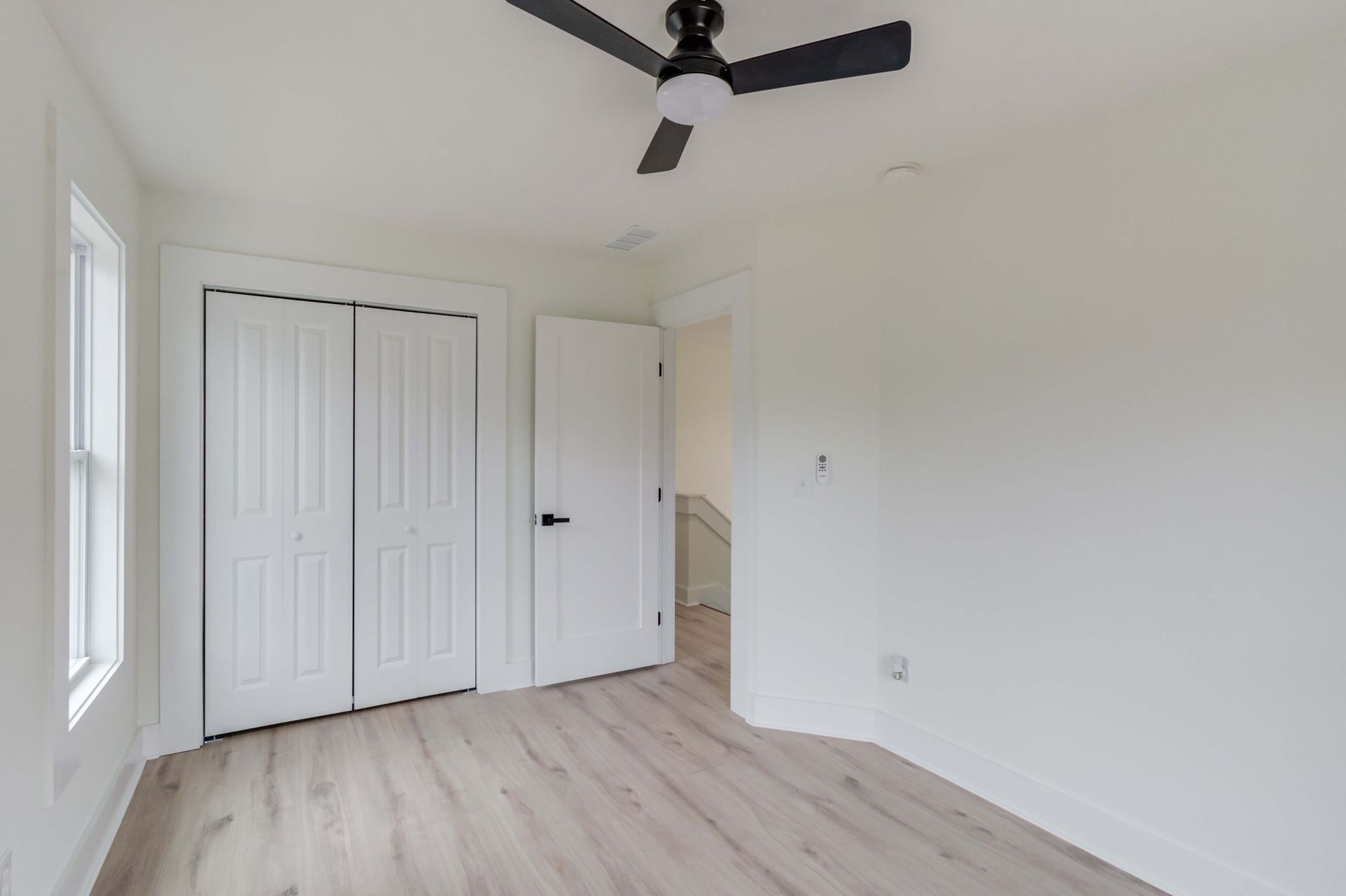 A brightly lit, empty bedroom with white walls, wood-look flooring, a ceiling fan, and white closet and room doors.