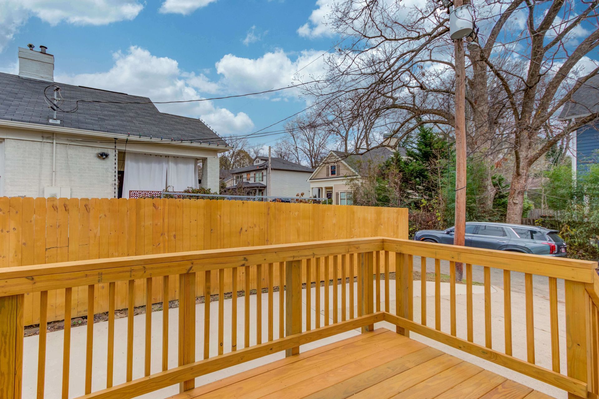 A wooden deck with railing overlooks a concrete yard and a wooden fence, with houses and trees in the background.