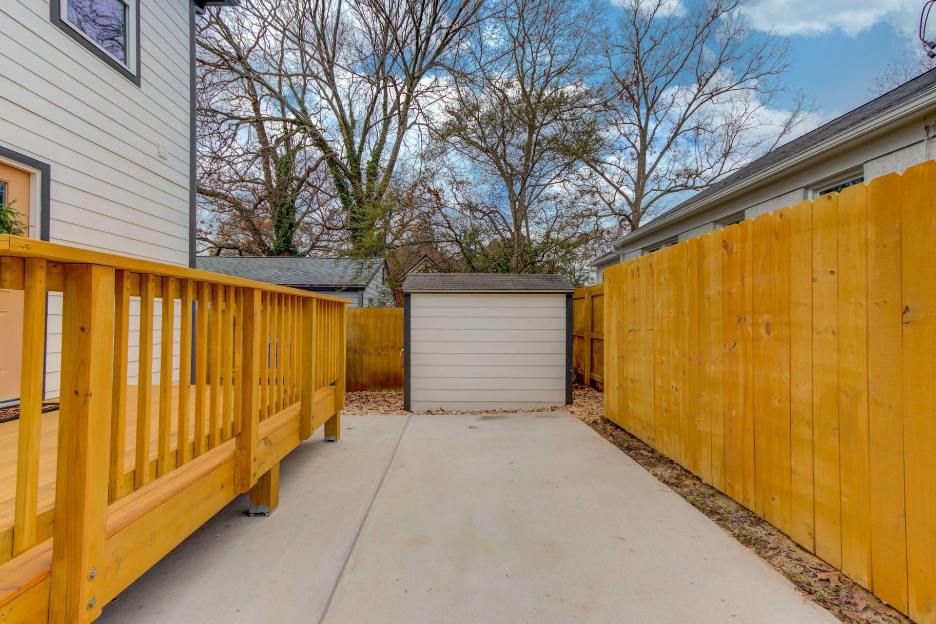 A concrete patio area between a wooden deck and a yellow fence, leading to a small shed under bare trees.