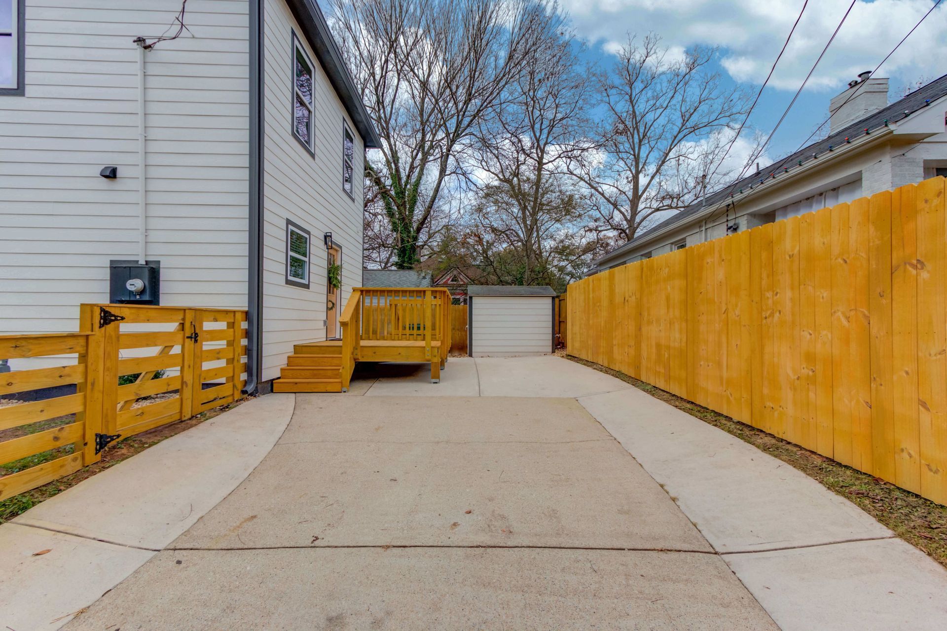 Concrete driveway leading to a garage, flanked by a wooden deck and fence alongside a light-colored house.