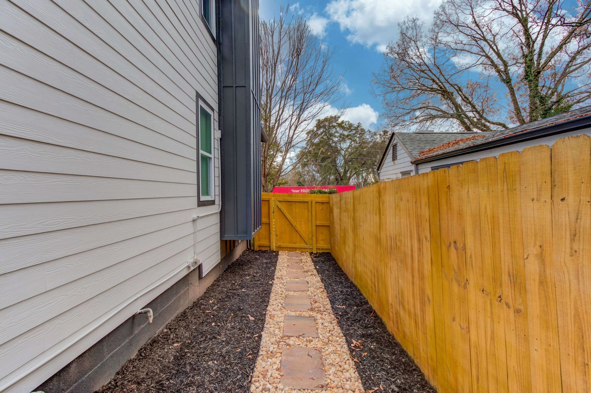 A narrow side yard with a gravel path, white siding on the left, and a wooden fence on the right leading to a yellow gate.