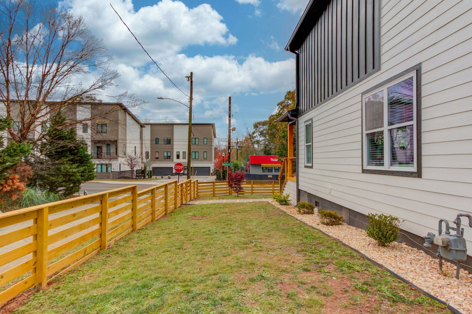 A grassy backyard with a wooden fence alongside the white exterior wall of a house under a partly cloudy sky.