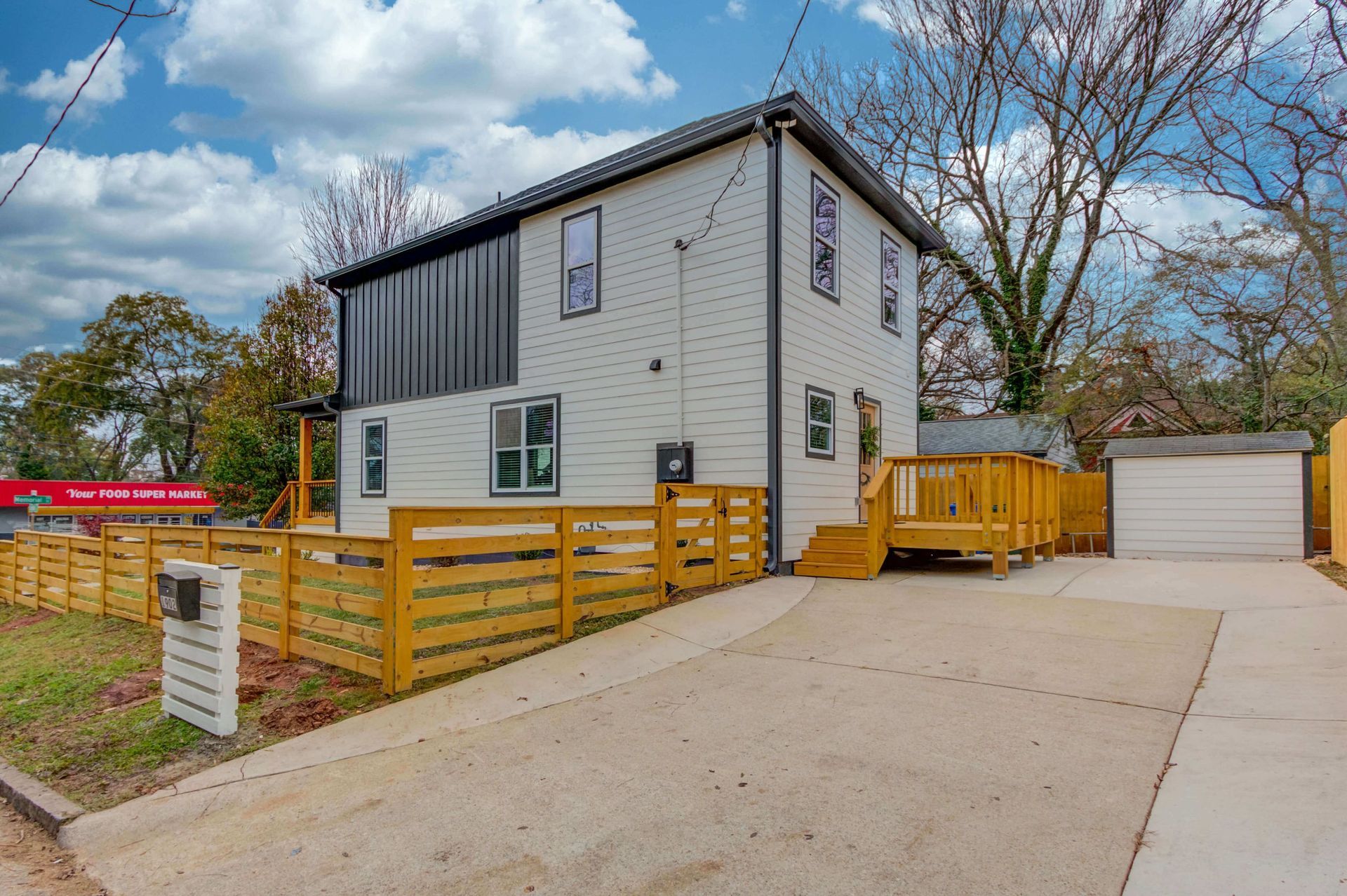 Two-story white house with a dark metal upper accent, wood fence, and concrete driveway under a cloudy blue sky.