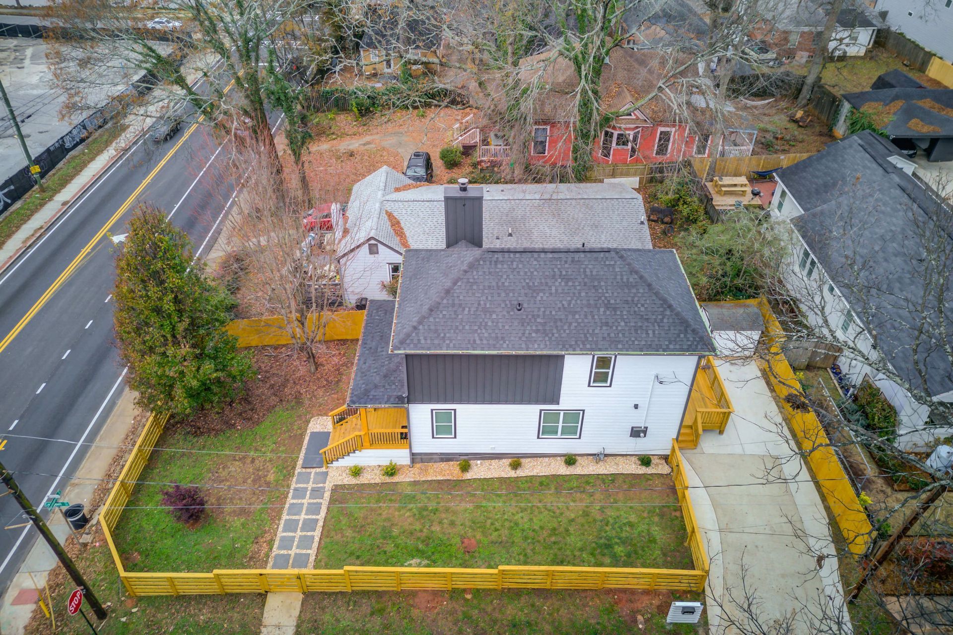 An aerial view of a white two-story house with a grey roof, enclosed by a yellow wooden fence next to a paved road.