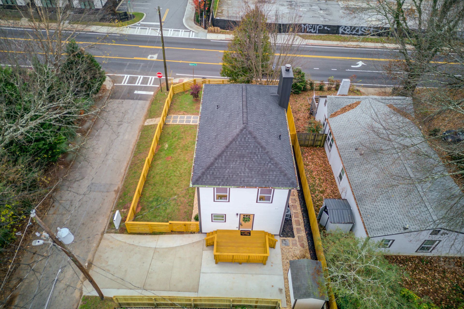 Aerial view of a white house with a dark gray roof, a fenced side yard, and a paved driveway next to a road.