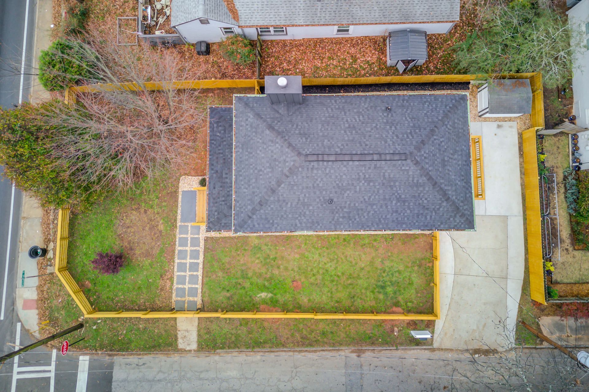 An aerial view of a single-story house with a dark gray shingled roof, a fenced front yard, and a concrete driveway.