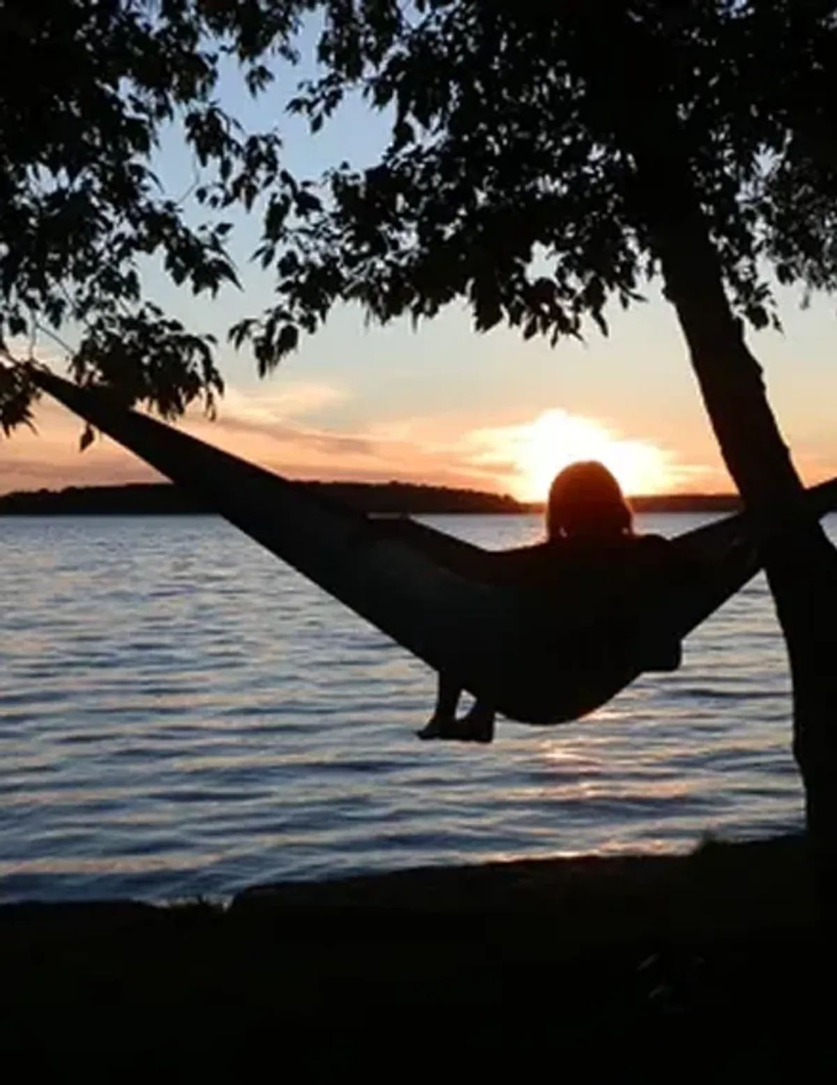 Person lounging in a hammock by the water at sunset, framed by trees.