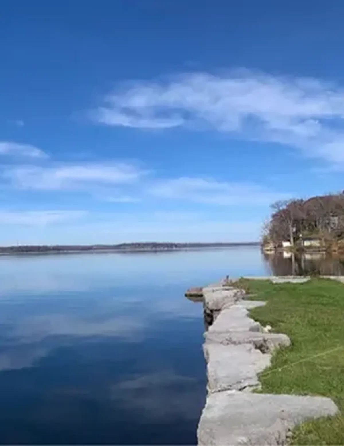 Calm lake under a blue sky, with a grassy shoreline and stone edge in the foreground