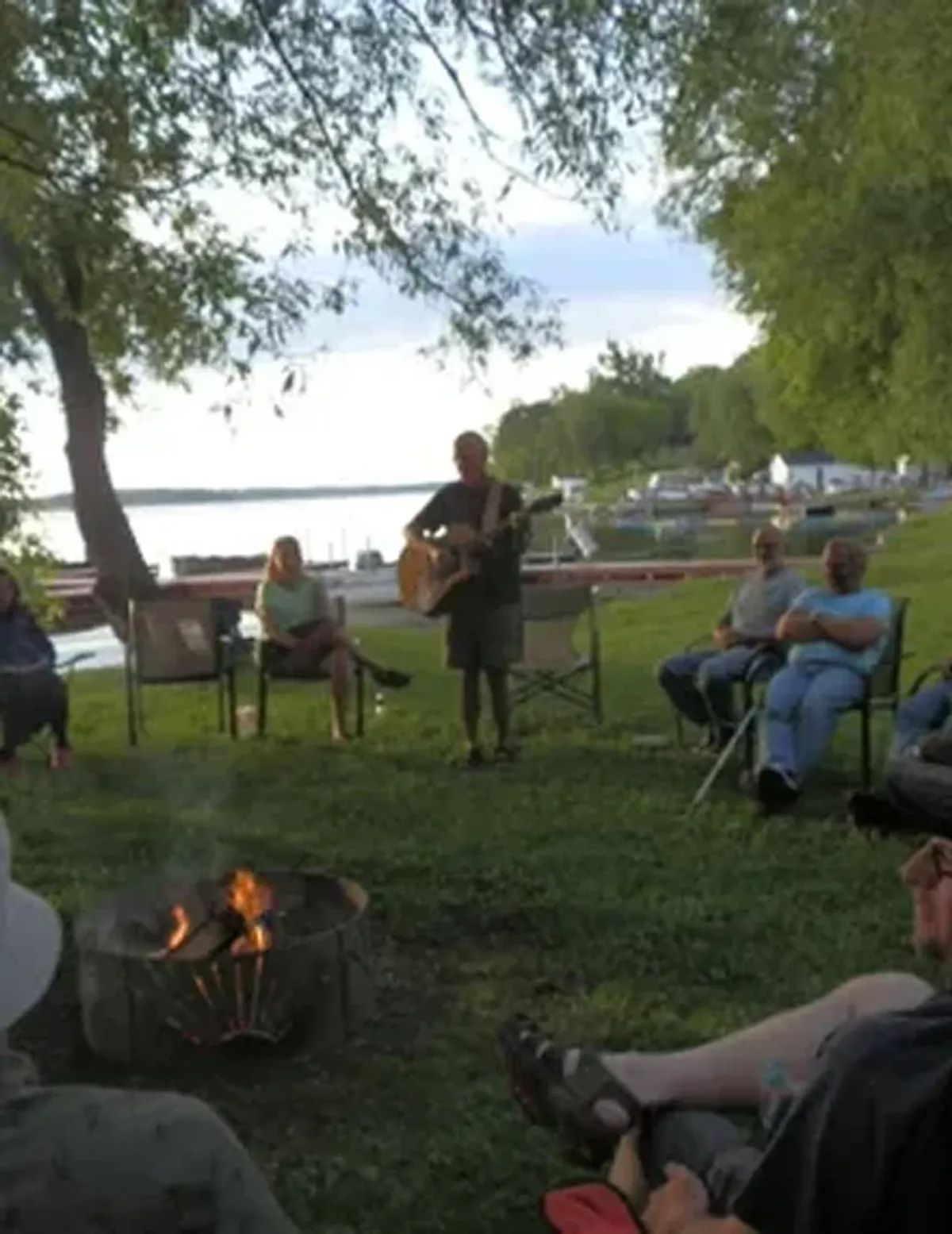 People sitting around a campfire under trees by a lakeside at dusk, one person playing guitar.