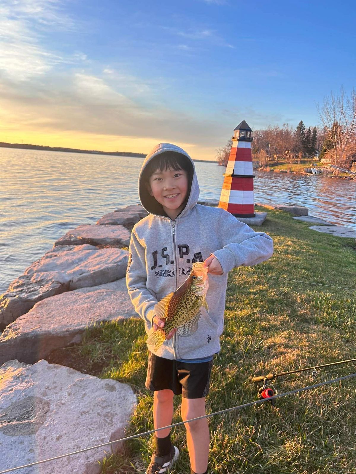 Child holding a fish by a rocky lakeshore at sunset, with a red-and-white lighthouse nearby
