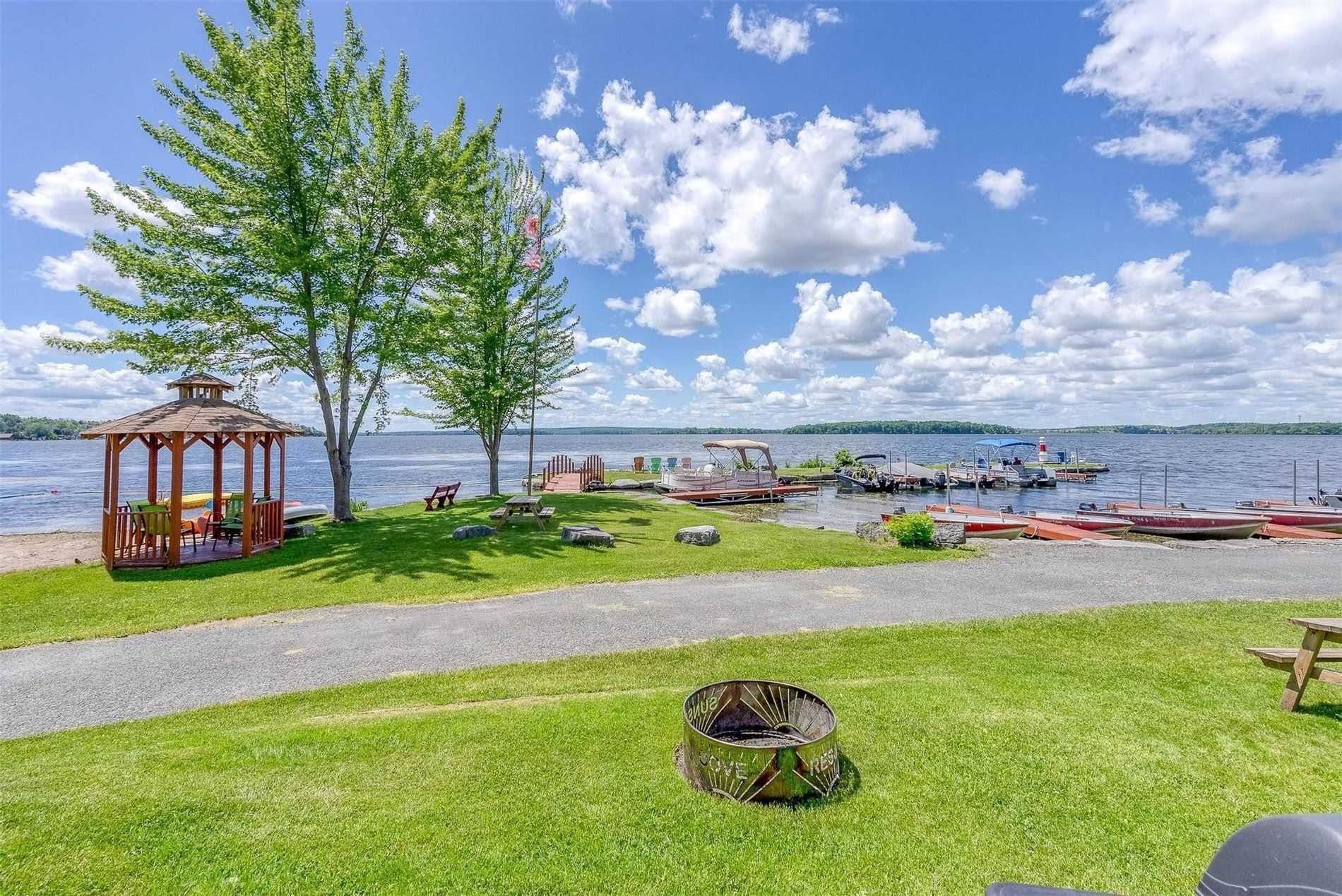Grassy lakeside park with a tree, gazebo, bench, and boats on a calm waterway under a blue sky