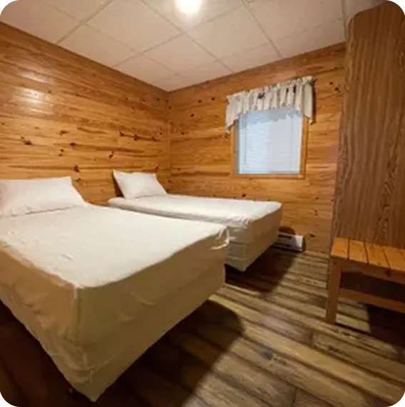 Small rustic bedroom with two white twin beds, wood-paneled walls, and a window with curtains.