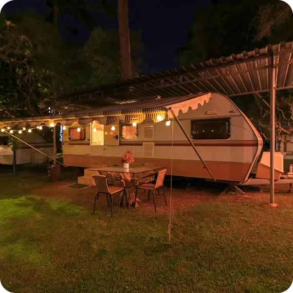 Lit vintage camper under string lights with outdoor table and chairs on a grassy campsite at night
