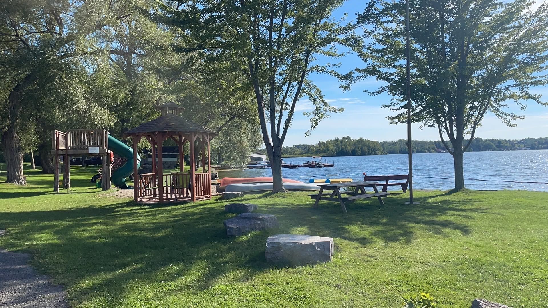 Lakeside park with picnic tables, playground, trees, and a rocky shoreline under a blue sky