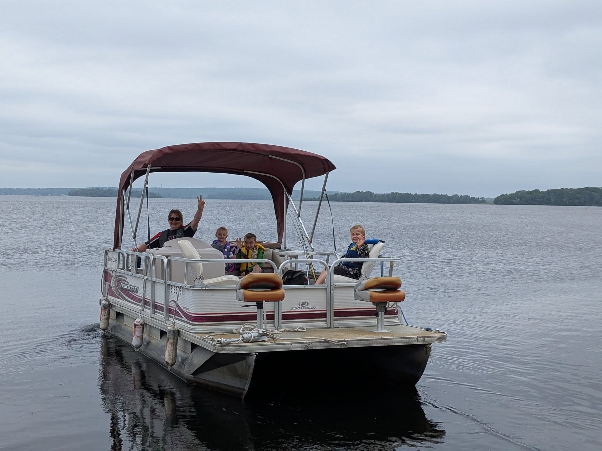 Pontoon boat with several people cruising on calm water under a cloudy sky