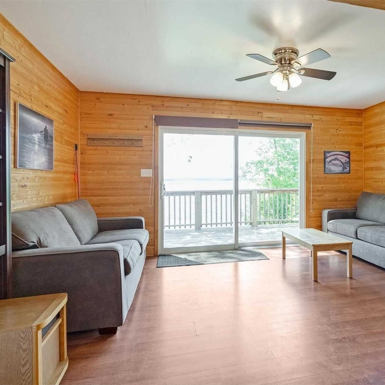 Cozy wood-paneled living room with gray sofas, coffee table, ceiling fan, and sliding glass doors to a deck