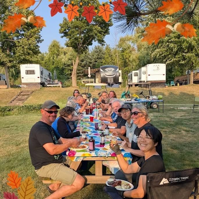 People sharing a long outdoor picnic table in a campground, with RVs and autumn trees in the background.