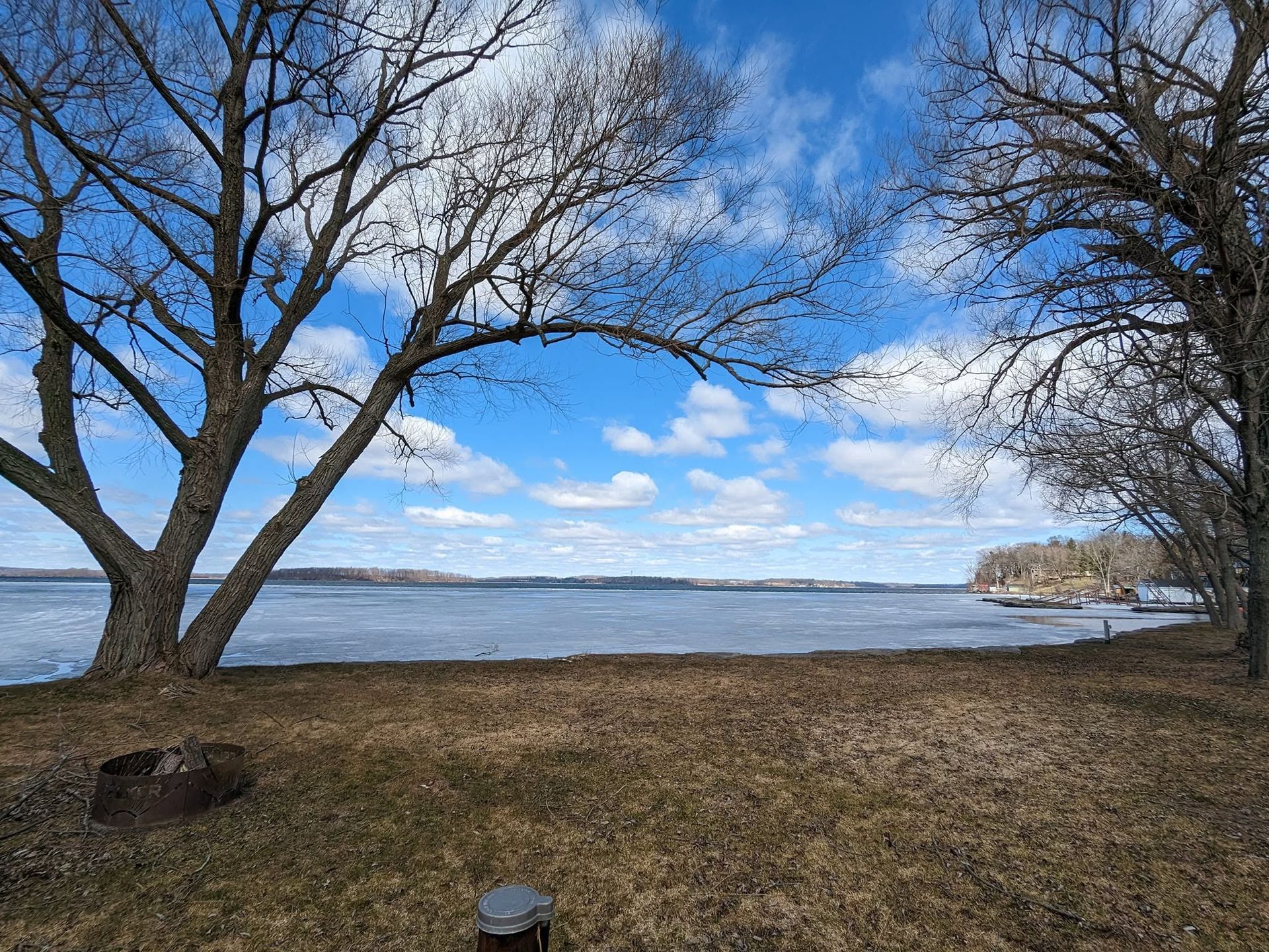 Leafless trees frame a calm lake under a bright blue sky with scattered clouds.