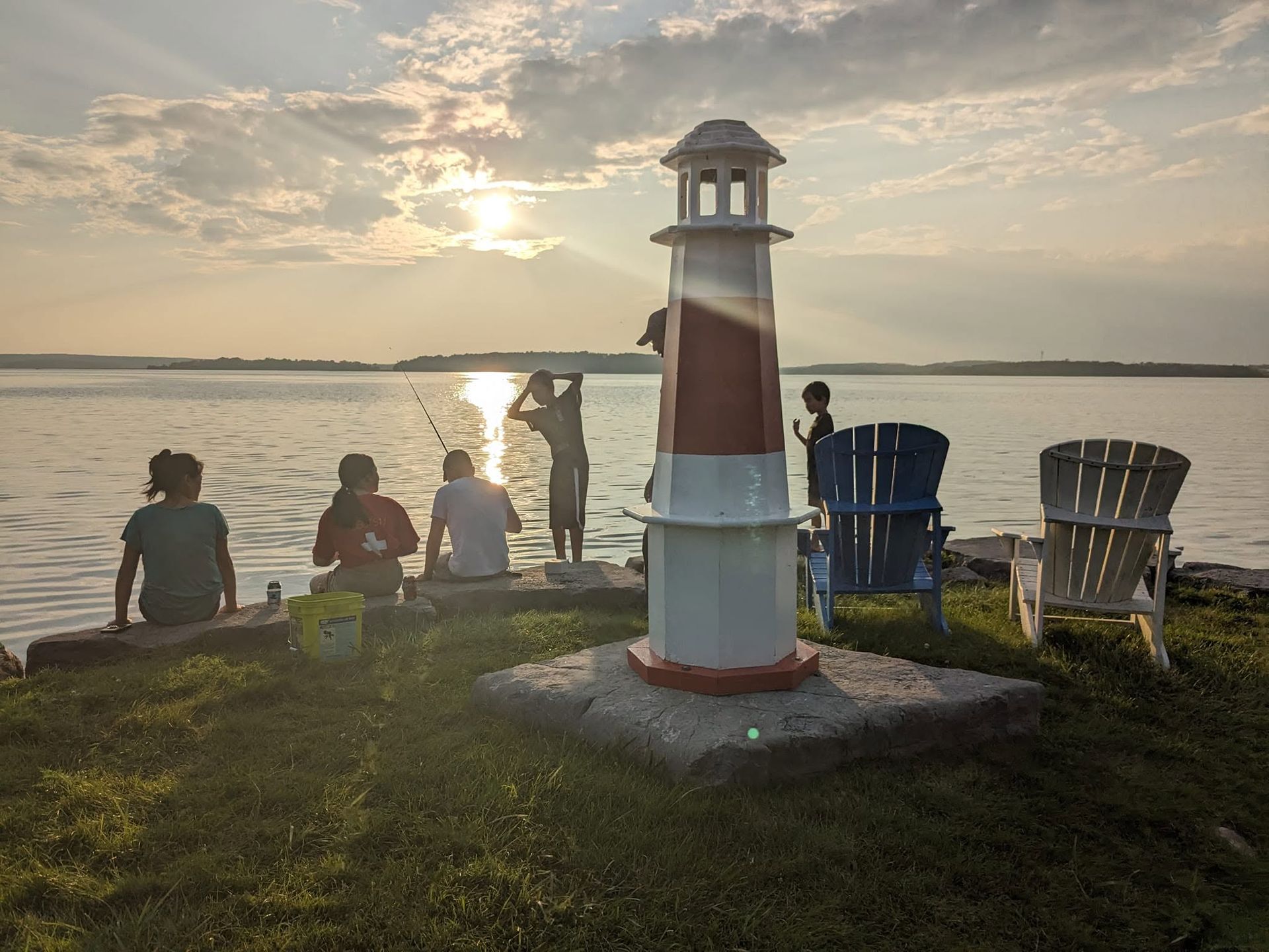People sitting by a lighthouse at sunset on a lakeshore, with colorful chairs and calm water.