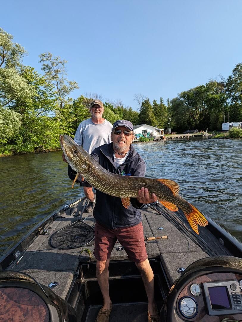 Angler holding a large fish on a dock, with another person behind and water in the background.