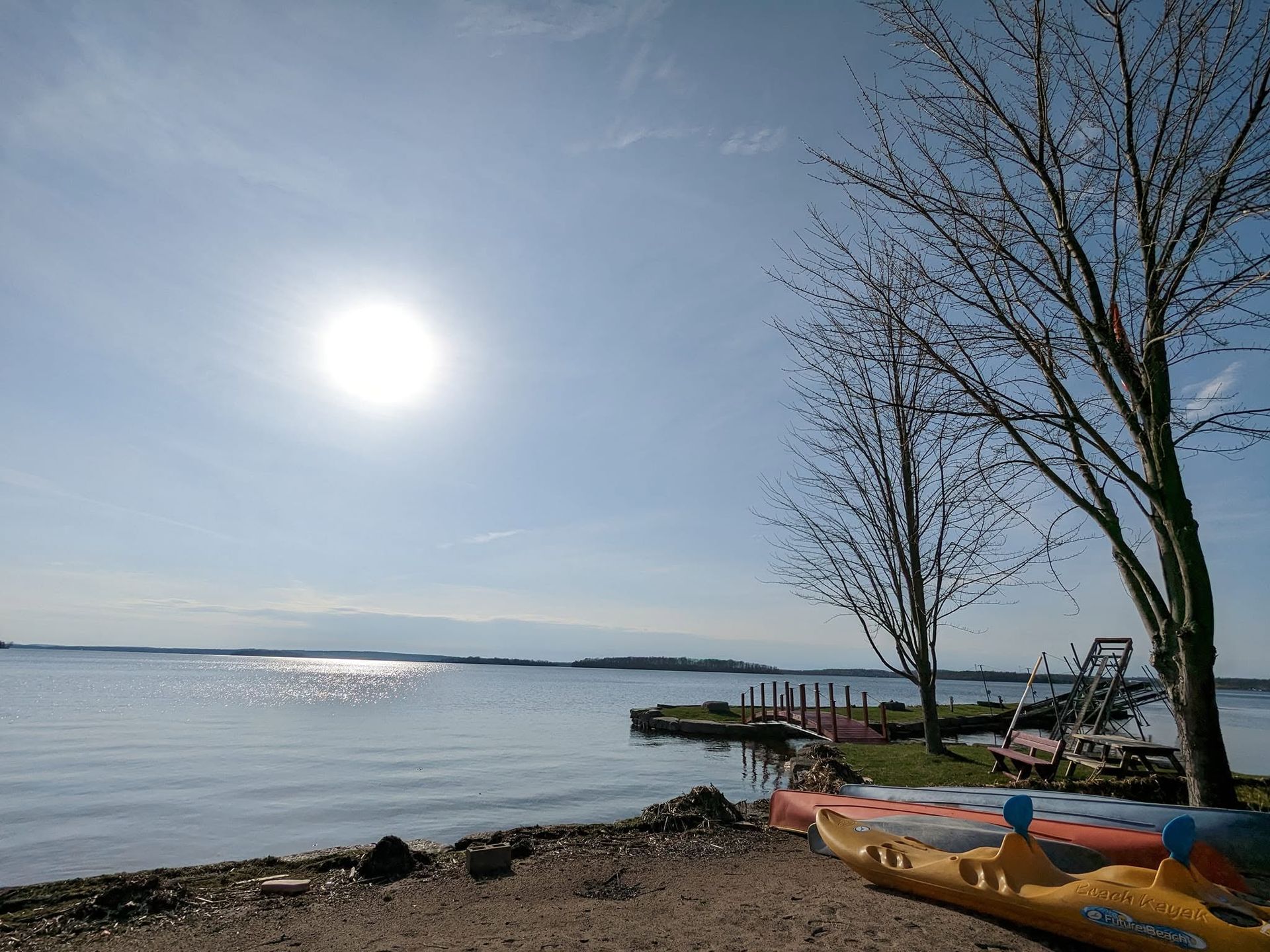 Sunlit lakeshore with calm water, dock, bare trees, and colorful kayaks on the shore