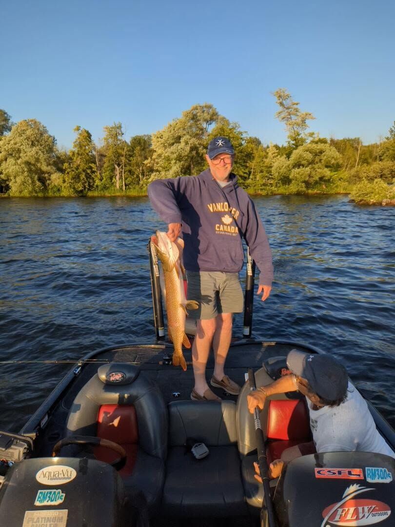 Person on a boat holding a large fish, with a dog nearby, on a lake at sunset.