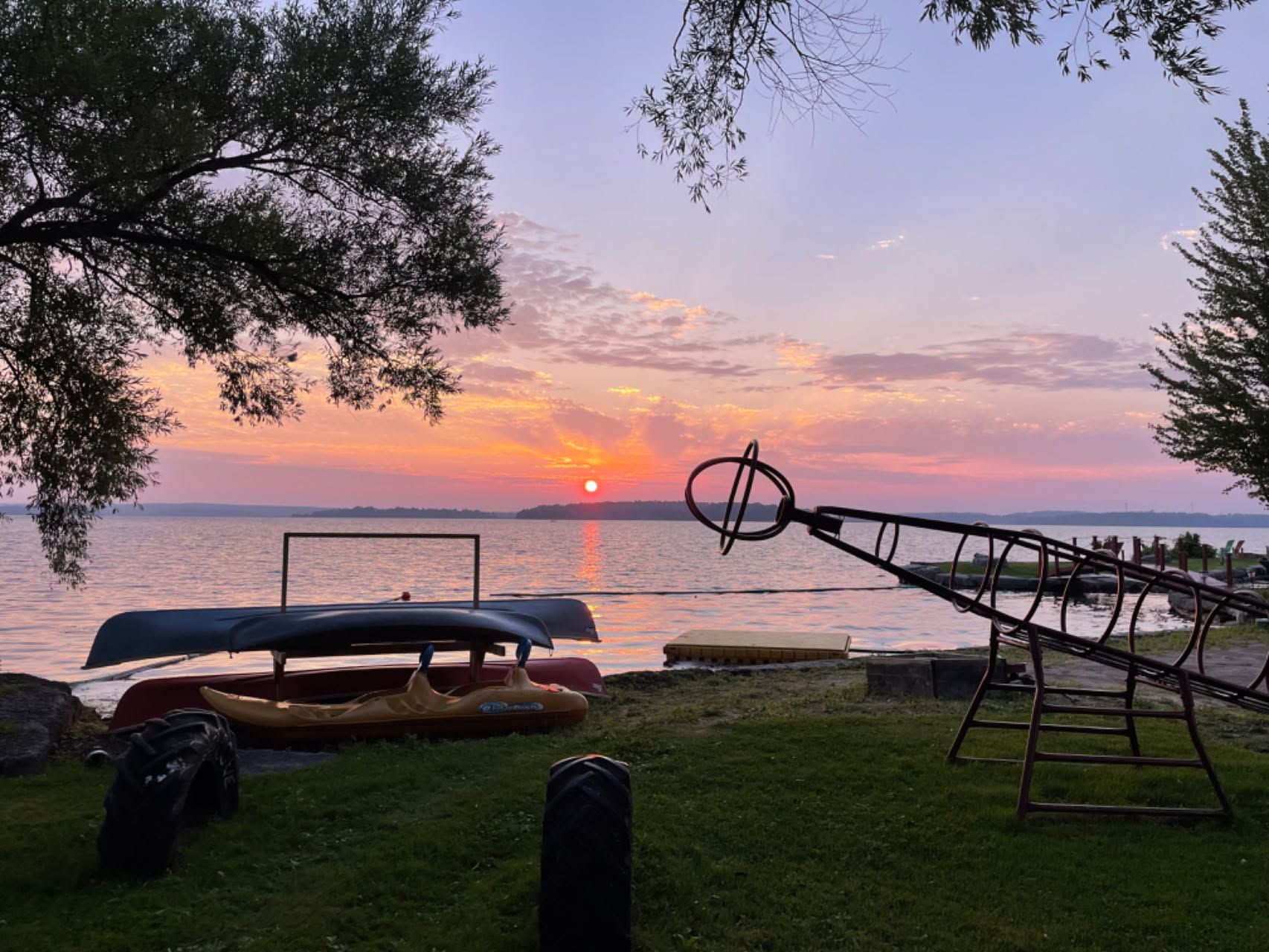Sunset over a lakeshore with dock equipment silhouetted on grassy shore and trees framing the view